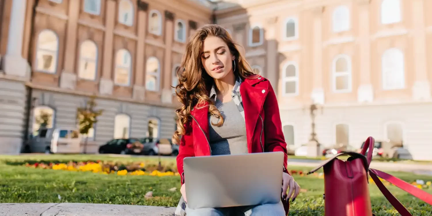 Outdoor portrait of serious curly female student sitting with laptop on the ground